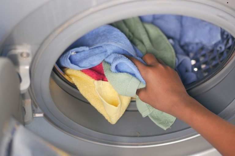 A hand reaching into a washing machine, holding colorful towels including yellow, green, red, and blue, ready for washing.