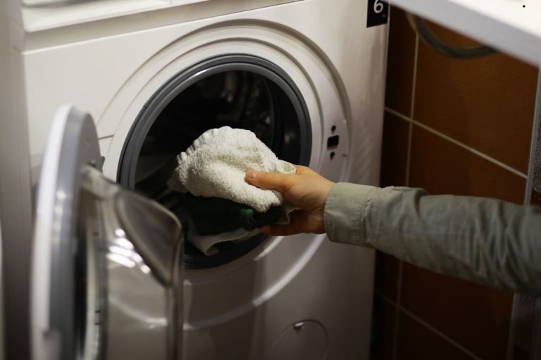 A hand placing a white towel into a front-loading washing machine. The machine is modern, set against tiled walls.